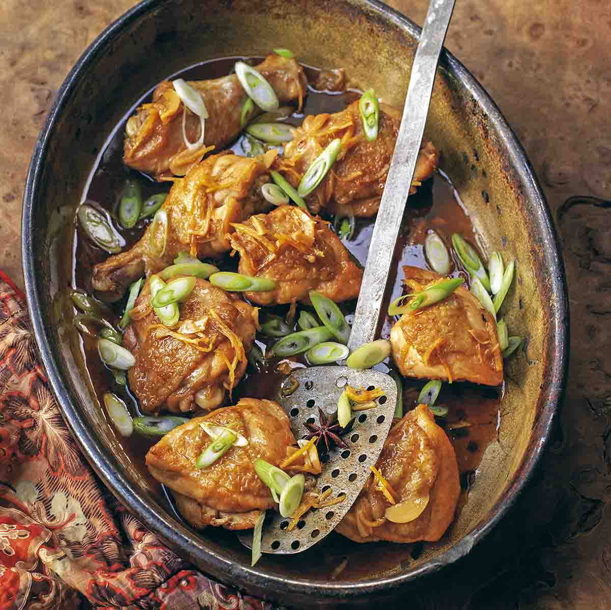 A crock filled with browned chicken thighs and sliced scallions, a spatula in the middle.
