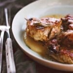 Chicken pieces in a sauce of vinegar, garlic, rosemary in a white plate, knife and fork on the side.