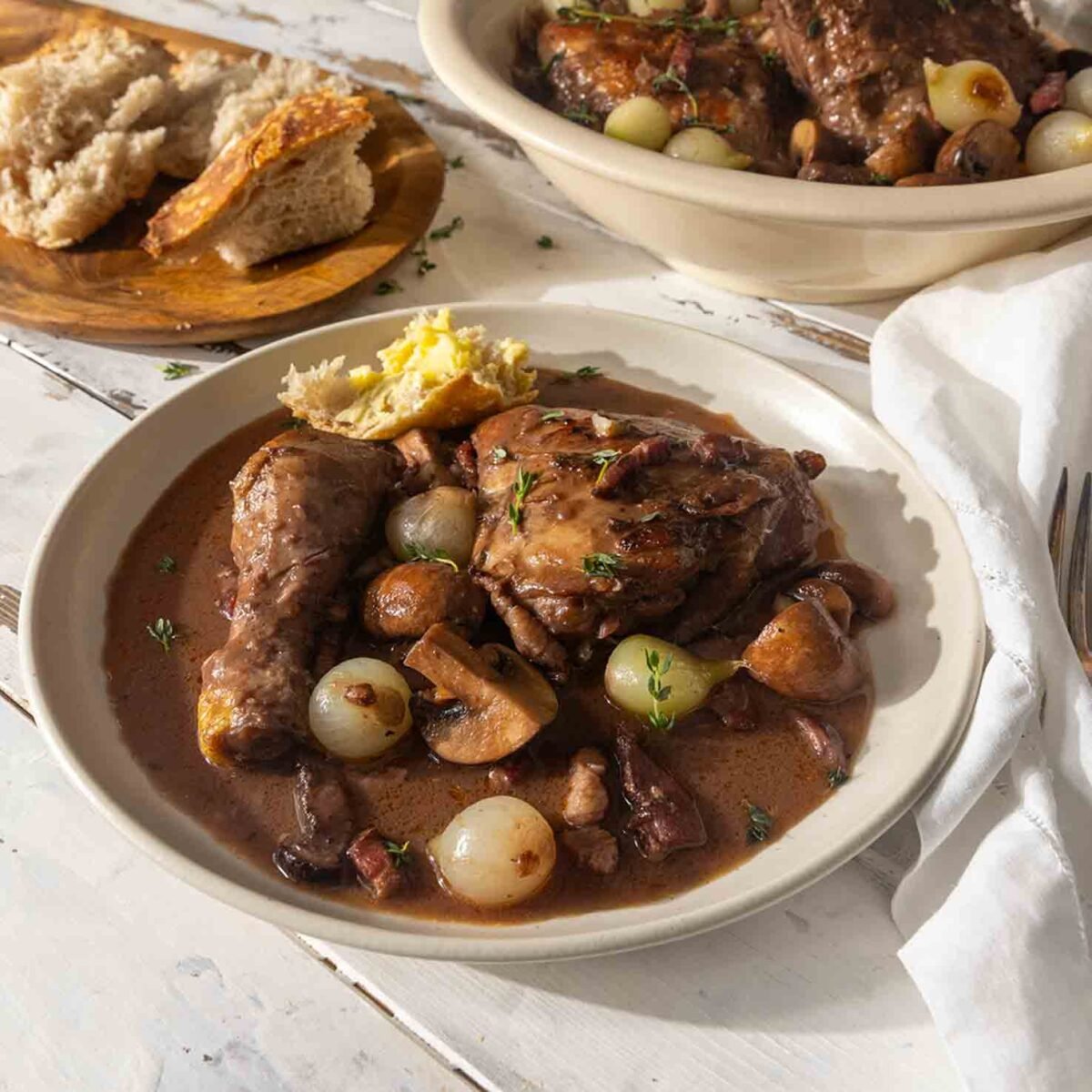 Two bowls of coq au vin on a table with sliced bread and fresh herbs.