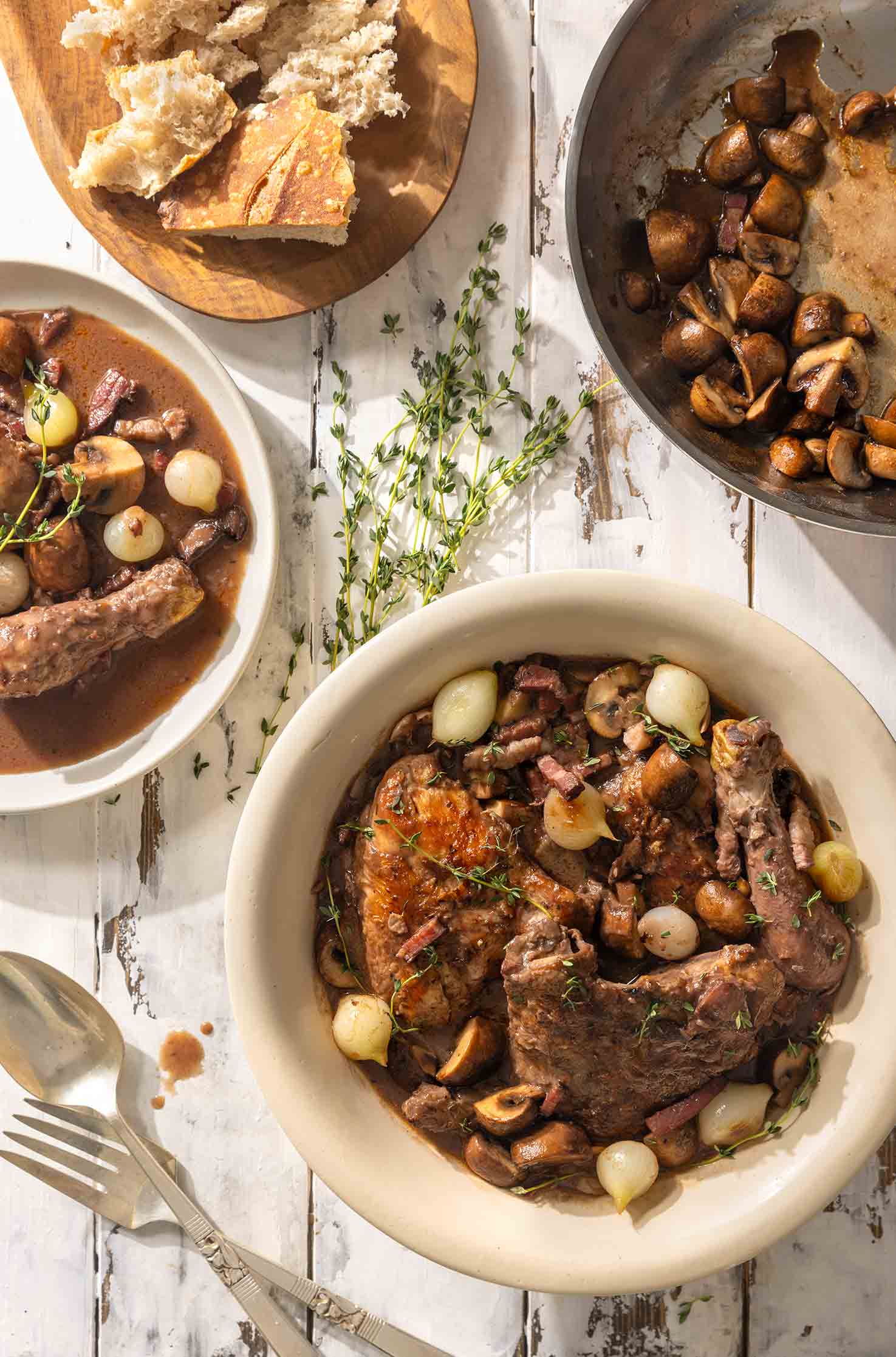 Two bowls of coq au vin on a table with sliced bread, sautéed mushrooms, and fresh herbs.