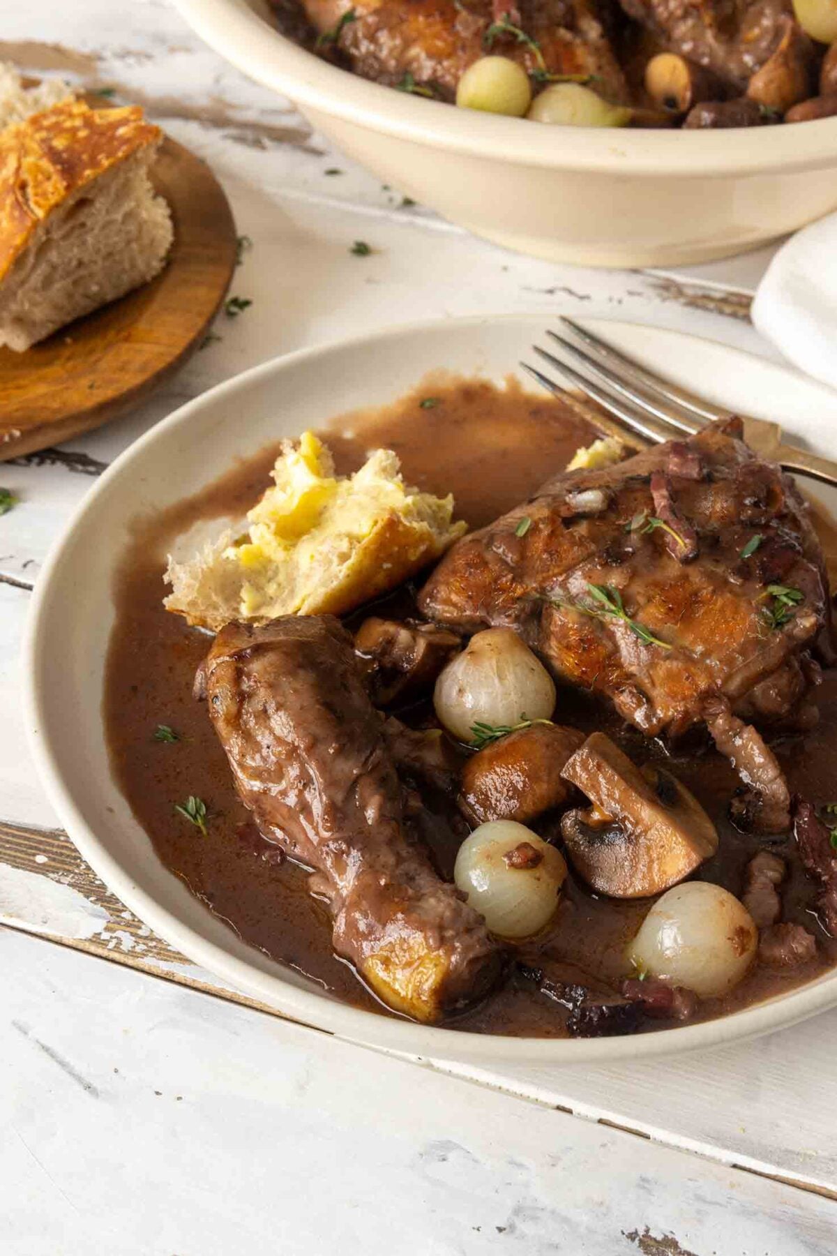 Two bowls of coq au vin on a table with sliced bread and fresh herbs.