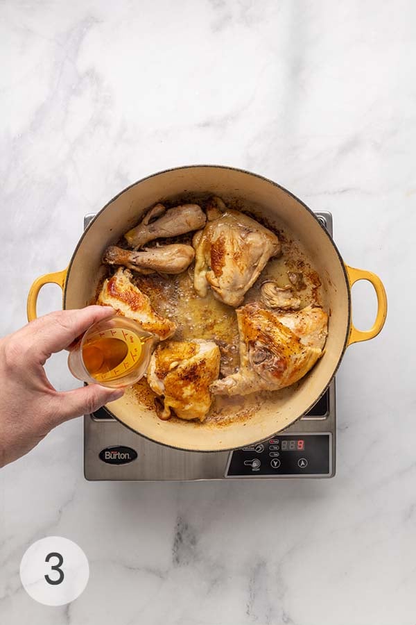 Cognac being poured into a Dutch oven with browned chicken pieces.