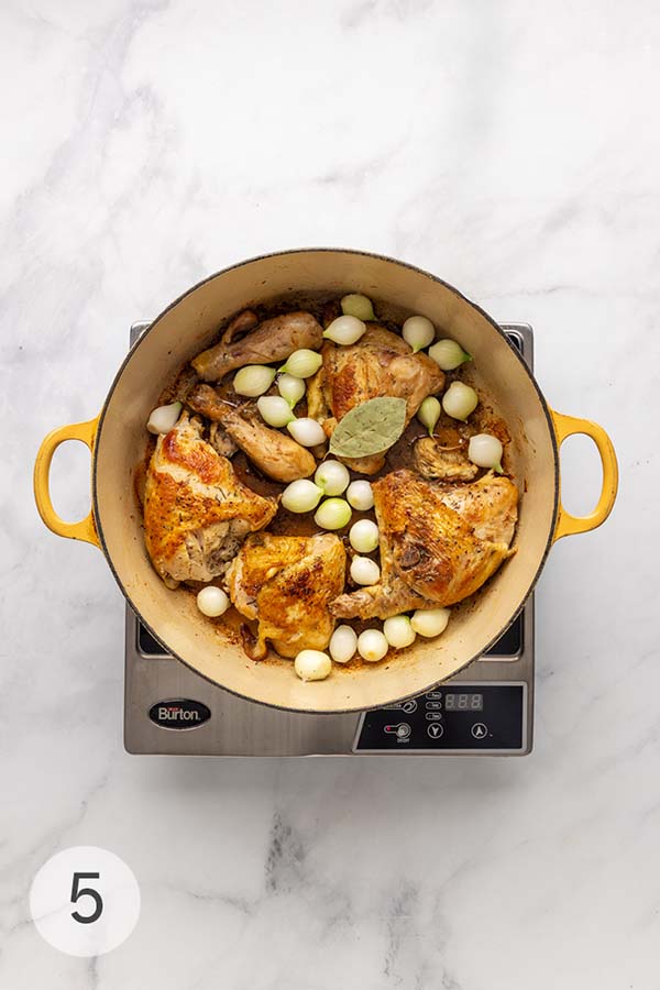 Browned chicken pieces in a Dutch oven with pearl onions and a bay leaf.