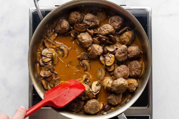 A person stirring meatballs and mushrooms in a skillet.