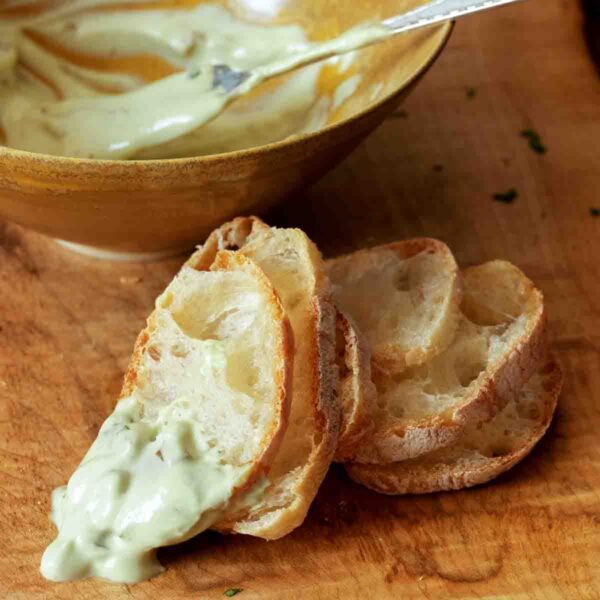 A bowl with creamy green olive dip behind five slices of baguette with dip on one.