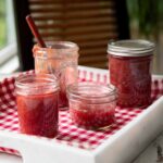 Four jars of rhubarb jam on a white tray lined with checkered cloth.
