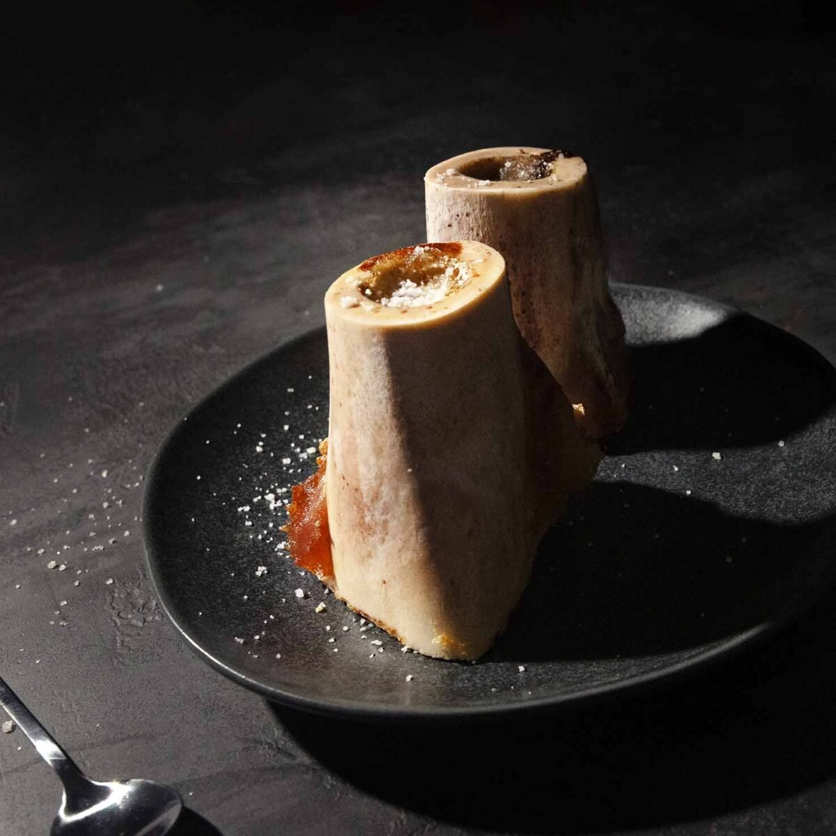 Two pieces of roasted bone marrow sprinkled with flaky salt on a black plate.