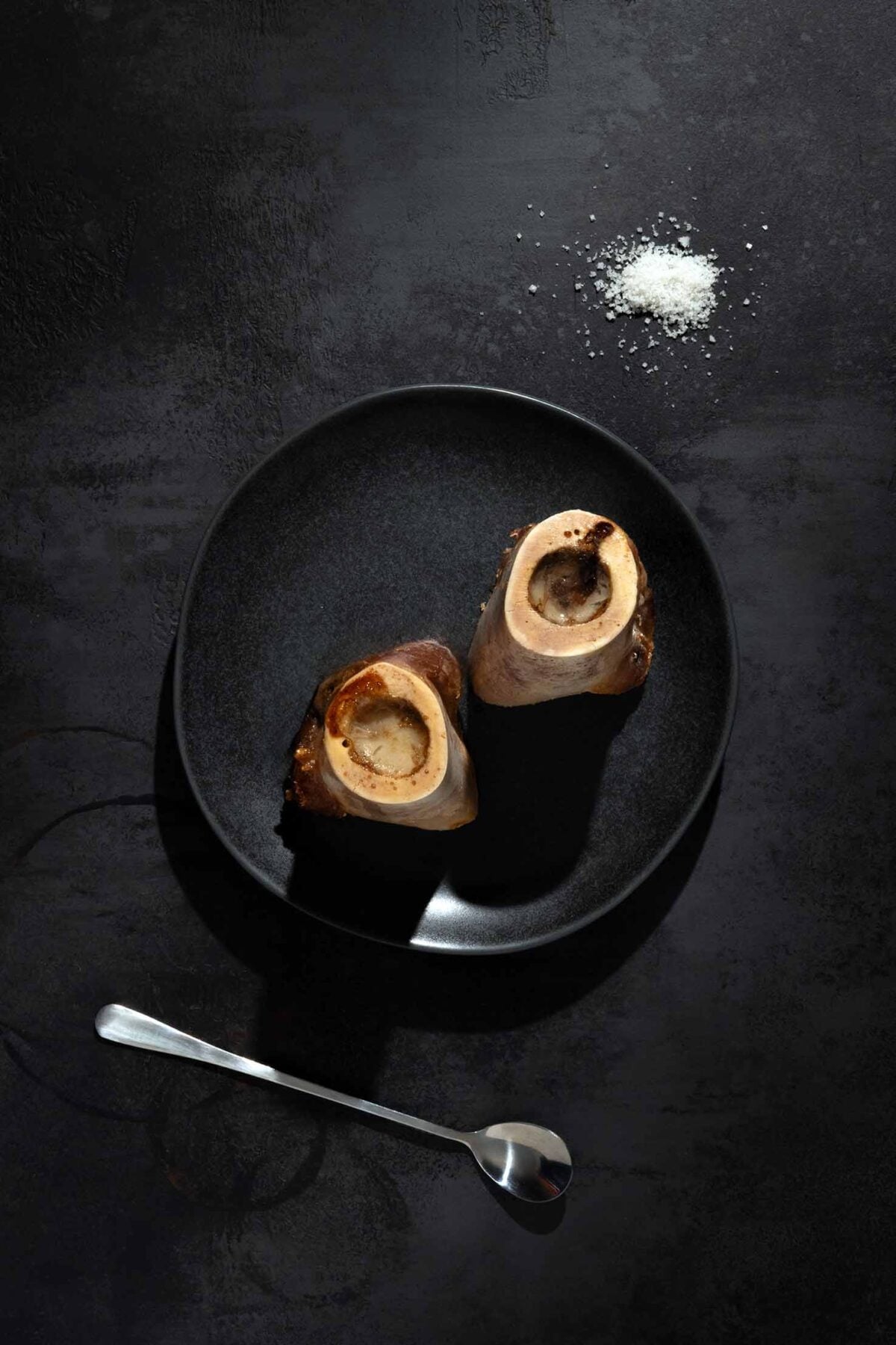 Two pieces of roasted bone marrow on a black plate with a pile of salt and a long handled spoon on the side.