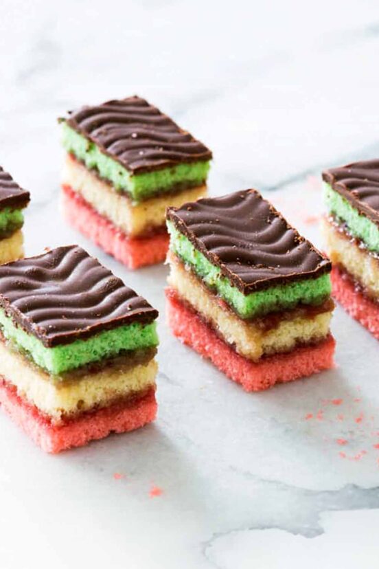 Several layered Italian rainbow cookies topped with chocolate on a marble surface.