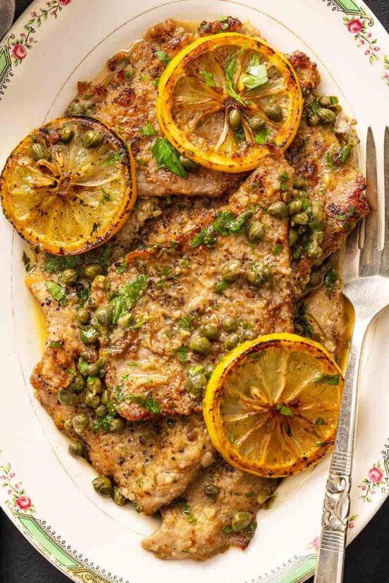 An oval platter of veal piccata--veal cutlets, lemon sauce, sliced lemons, capers, and chopped parsley.