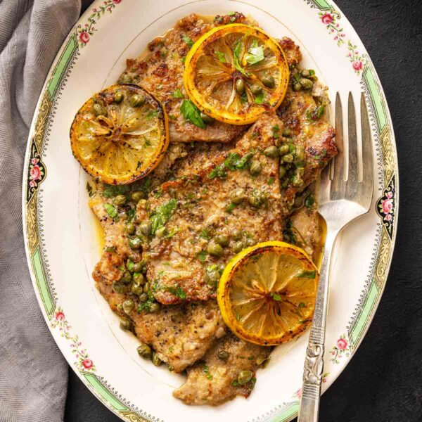 An oval platter of veal piccata--veal cutlets, lemon sauce, sliced lemons, capers, and chopped parsley.