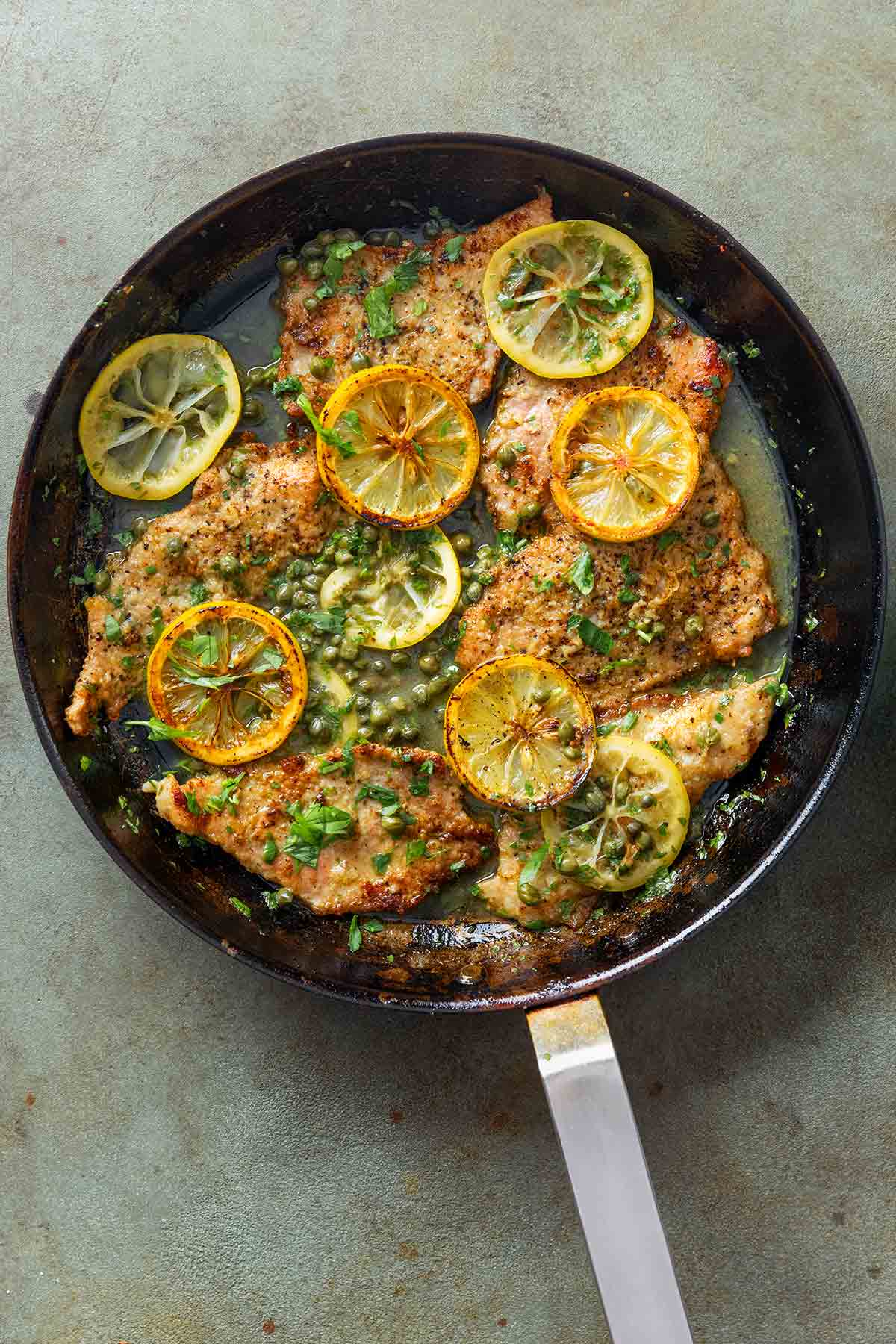 A black skillet of veal piccata--veal cutlets, lemon sauce, sliced lemons, capers, and chopped parsley.
