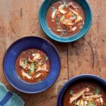 Three blue bowls filled with ancho chile soup on a wooden table.