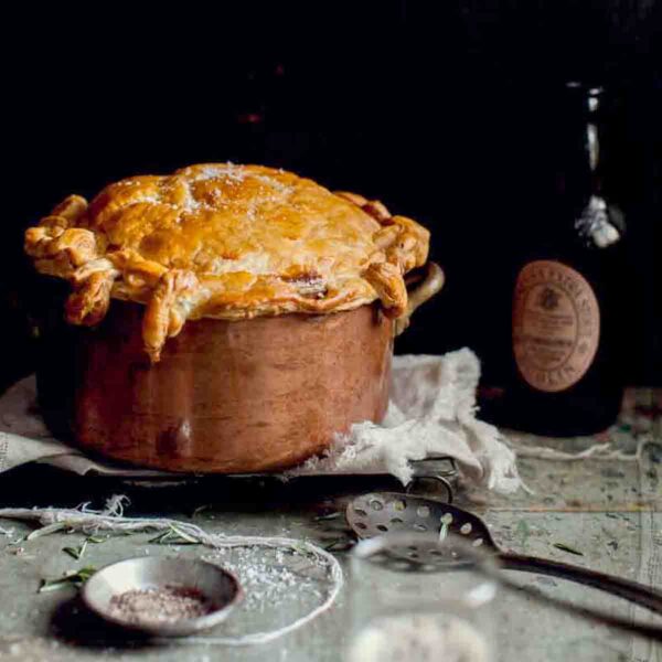 A copper pot filled with beef and Guinness pie topped with puff pastry on a towel on a cooking rack with a bottle behind it.