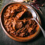 An oval bowl filled with braised pork in red chile sauce with a spoon resting on the side and a stack of tortillas and dried peppers on the side.