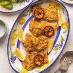 Three breaded chicken thighs on a plate with slices of roasted lemon.