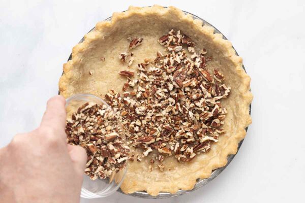 A person pouring toasted pecans into a baked pie crust.