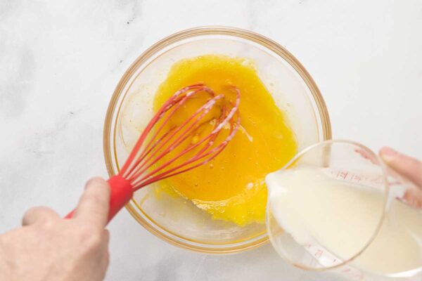 A man's hand whisking hot milk into egg yolks.