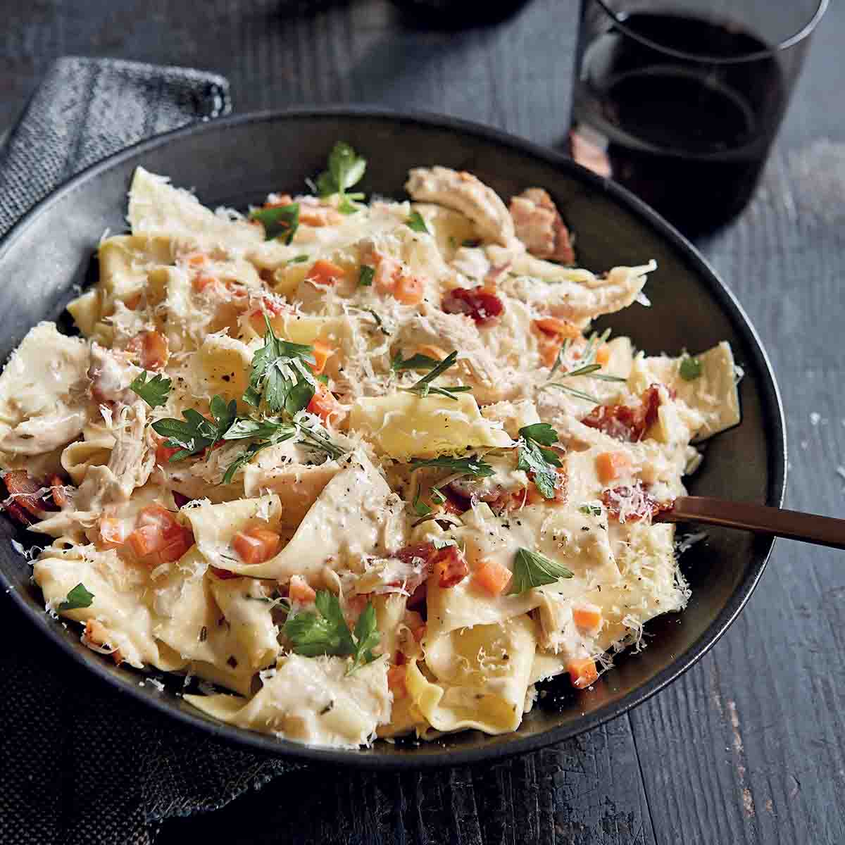 Creamy pappardelle with chicken and bacon in a black bowl with a fork, topped with grated cheese.