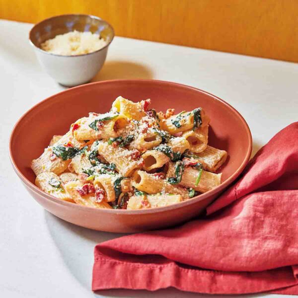 A red bowl filled with creamy sun-dried tomato and spinach pasta with a red cloth napkin on the side.