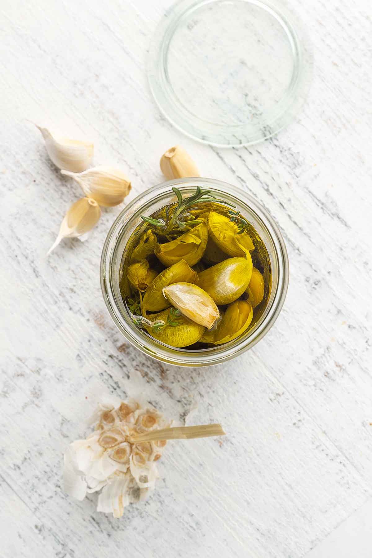 Garlic confit cloves in a jar of oil with some loose cloves nearby.