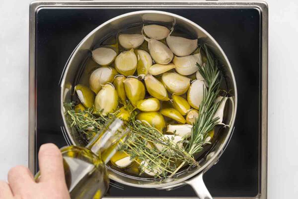 A person adding oil to a pot of garlic, thyme, and rosemary.