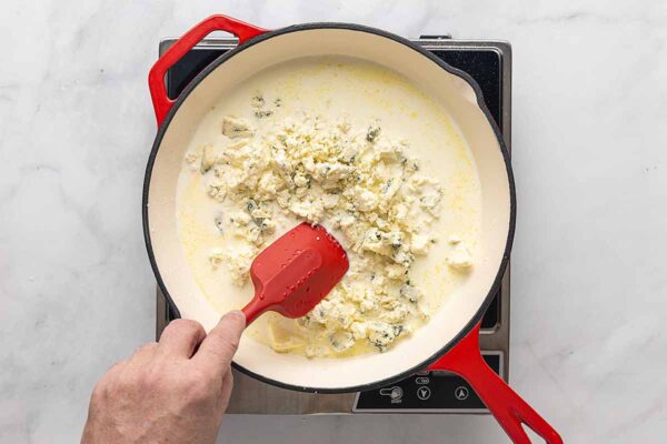 A person stirring crumbled blue cheese into cream in a skillet.