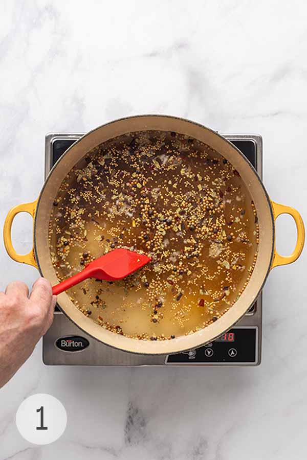 A pot of corned beef brine simmering with mustard seeds, peppercorns, and spices, stirred with a red spatula.