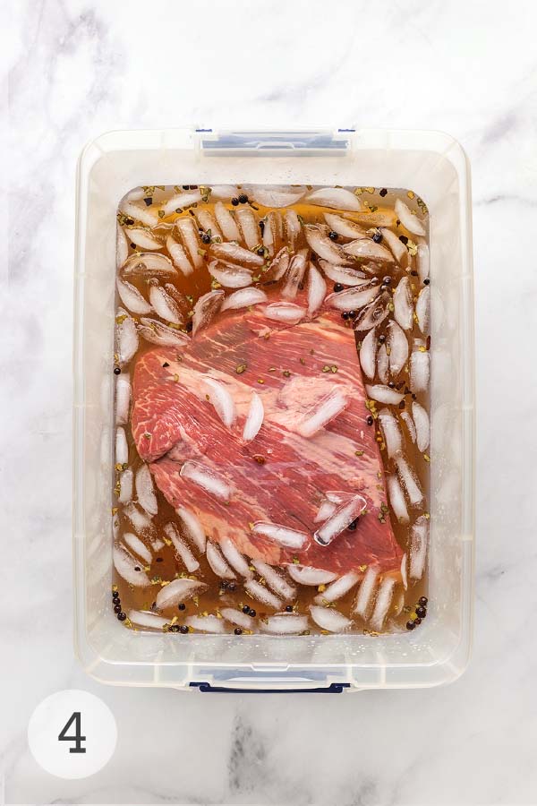 A raw brisket submerged in an ice-cold corned beef brine with peppercorns and spices floating on top.