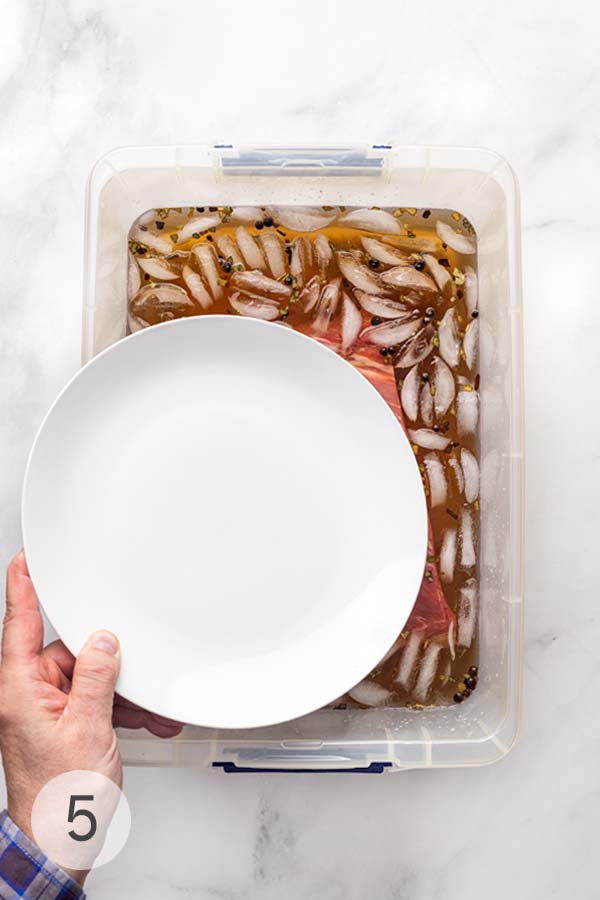 A hand placing a white plate on top of a brisket in a brine container to keep it submerged.