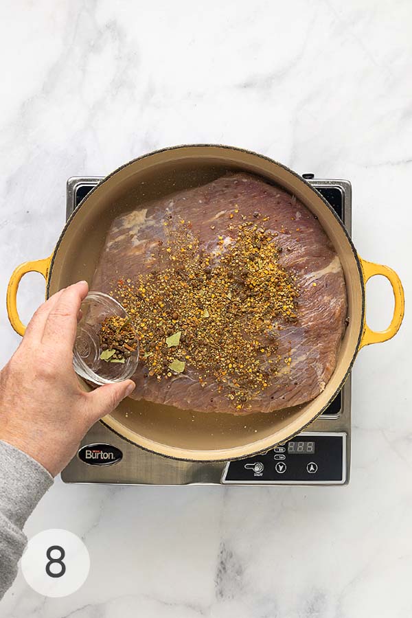 A hand adding a spice mix to a Dutch oven with corned beef submerged in water.