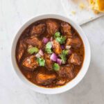 A white bowl filled with New Mexican red pork chili, garnished with red onion and cilantro.