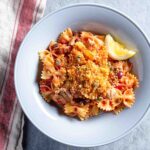 A pale blue bowl filled with farfalle, tuna, capers, tomatoes, and topped with bread crumbs and a slice of lemon, sitting on a red and white dish cloth.