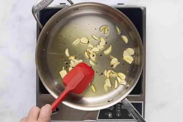 A person cooking garlic slices and pepper flakes in a skillet.
