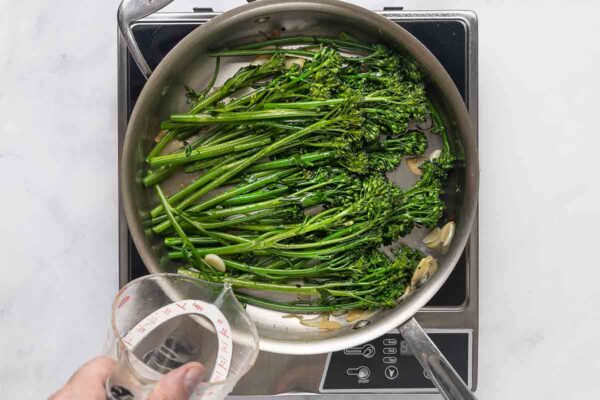 Broccolini and garlic in a skillet with a person pouring liquid into the skillet.