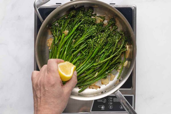 A person squeezing a lemon half into a skillet filled with broccolini and garlic.