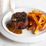 A person drizzling sauce over a steak au poivre on a white plate with sweet potato fries.