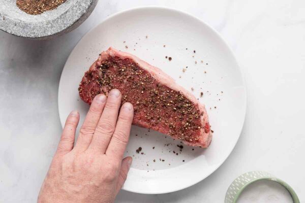 A person pressing a peppercorn mixture into a steak on a white plate.
