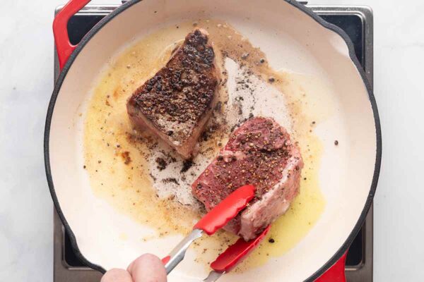 A person flipping seared steaks in a cast iron skillet.