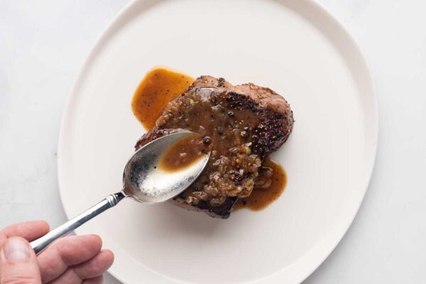 A person drizzling poivre sauce over a seared steak on a white plate.