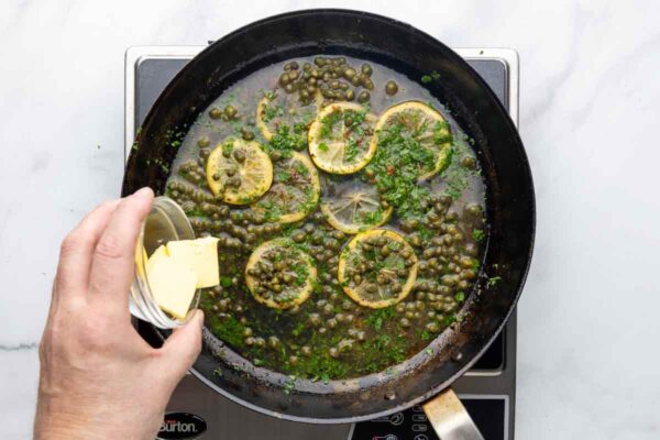 Butter being added to a skillet with lemon slices, parsley, and capers.