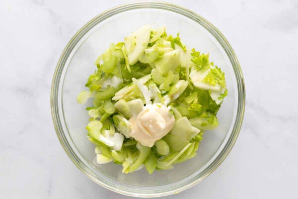 Sliced celery, celery leaves, and apple slices in a glass bowl with a drizzle of cream and dollop of mayonnaise on top.