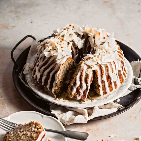 A banana coconut rum bundt cake on a metal try with a slice cut from in on a plate nearby.