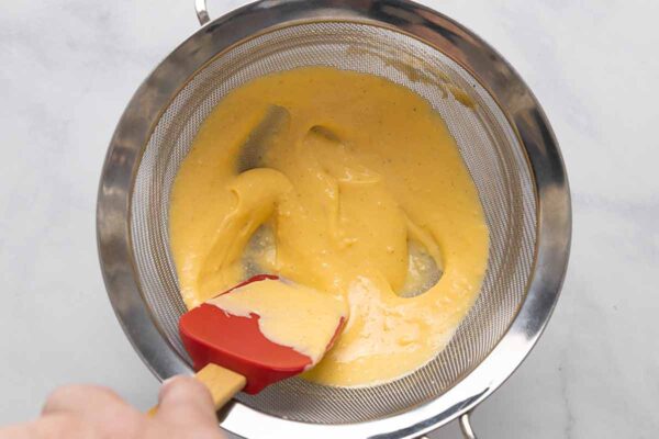 Cheese sauce being strained through a fine mesh sieve.