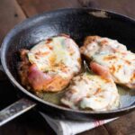 Three chicken cutlets wrapped in prosciutto and covered in cheese in a skillet.