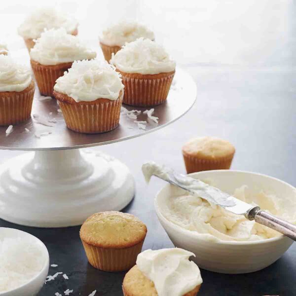 Cake stand with 7 frosted coconut cupcakes, four unfrosted cupcakes on the table, and a bowl of frosting.