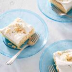 Three pieces of white cake topped with frosting and toasted coconut on individual blue glass plates with forks resting on the side.