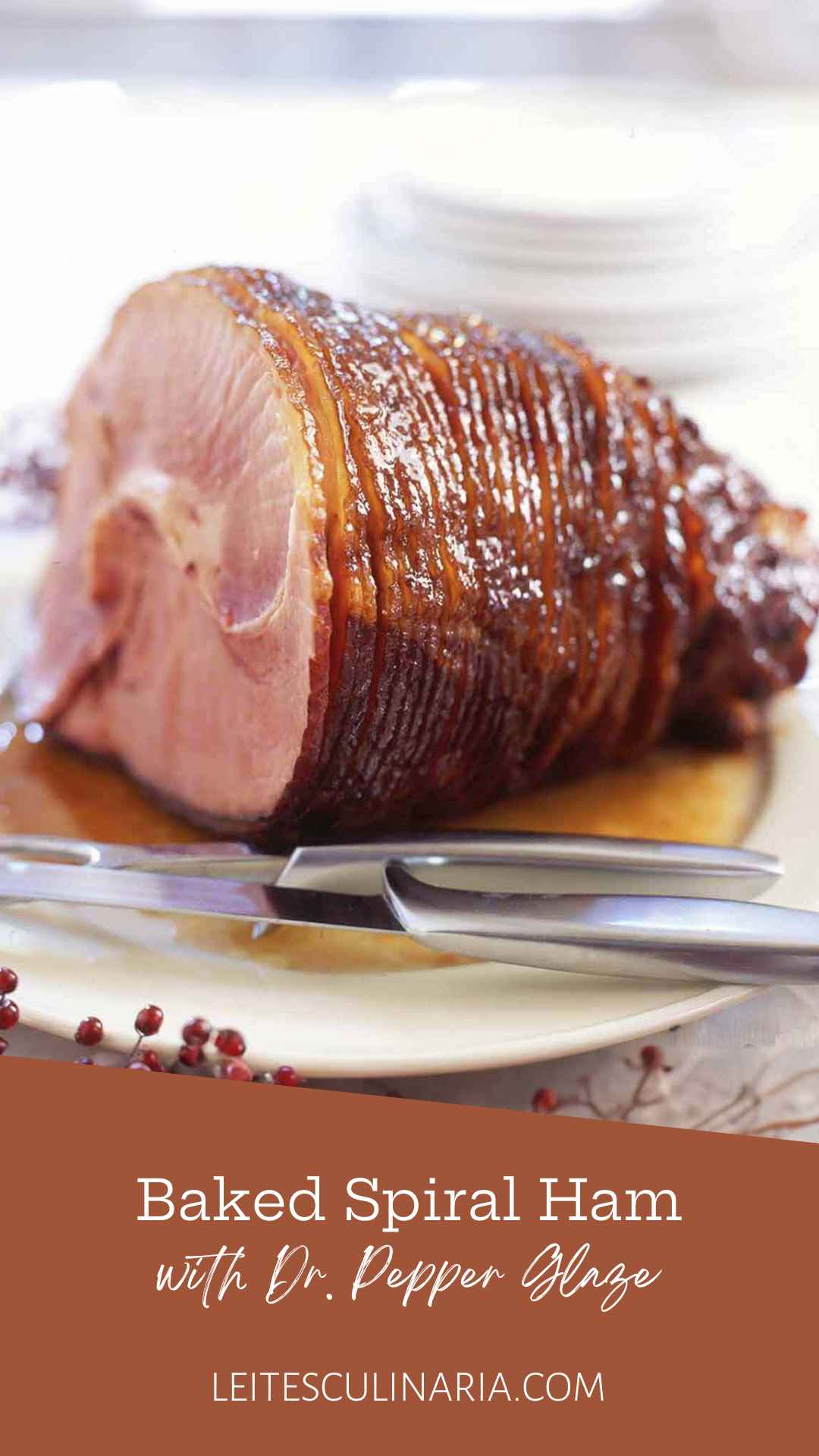 A glazed spiral cut ham on a white platter with a carving knife and fork nearby.