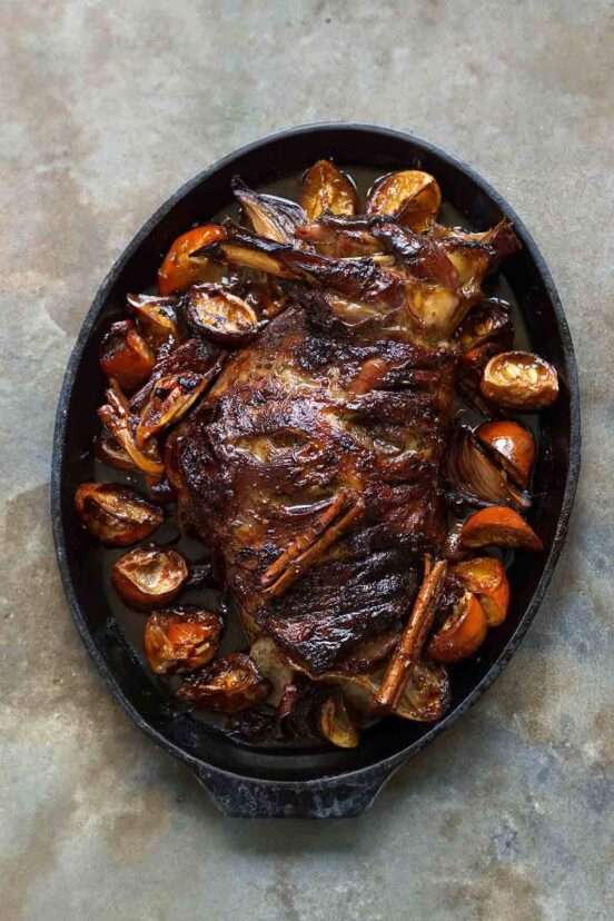 A slow roasted leg of lamb surrounded by orange segments and cinnamon sticks.
