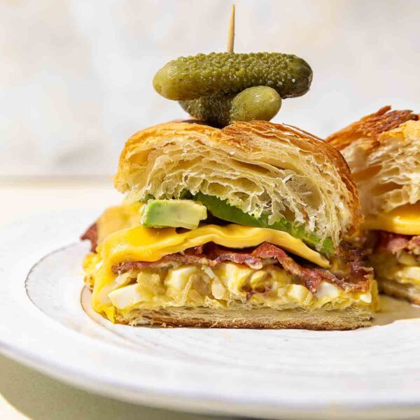 A halved croissant sandwich with egg salad, bacon, cheese, avocado, and small pickles on a white plate.